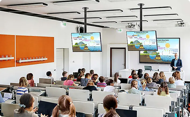 Lecture hall filled with students watching a presenter at the front; four screens display the presentation surface.