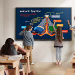 Teacher and three students point to a colorful volcanic eruption diagram on a large touch screen in a bright classroom.
