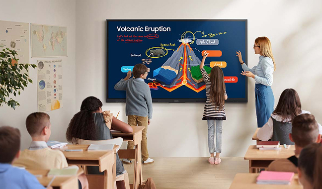Teacher and three students point to a colorful volcanic eruption diagram on a large touch screen in a bright classroom.