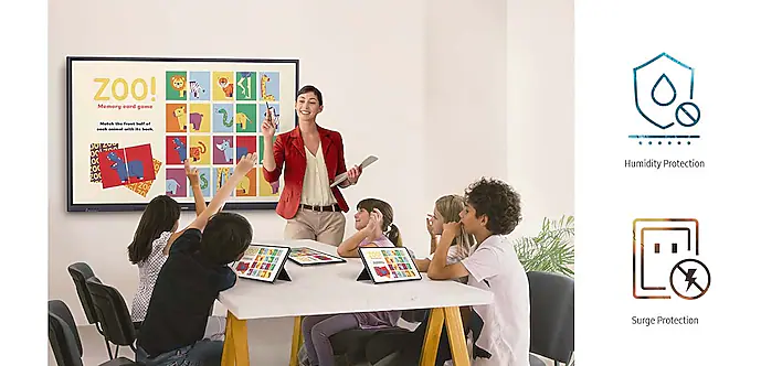 Teacher in a red blazer leads a classroom workshop with students around a table using tablets and a colorful poster on the wall behind them.
