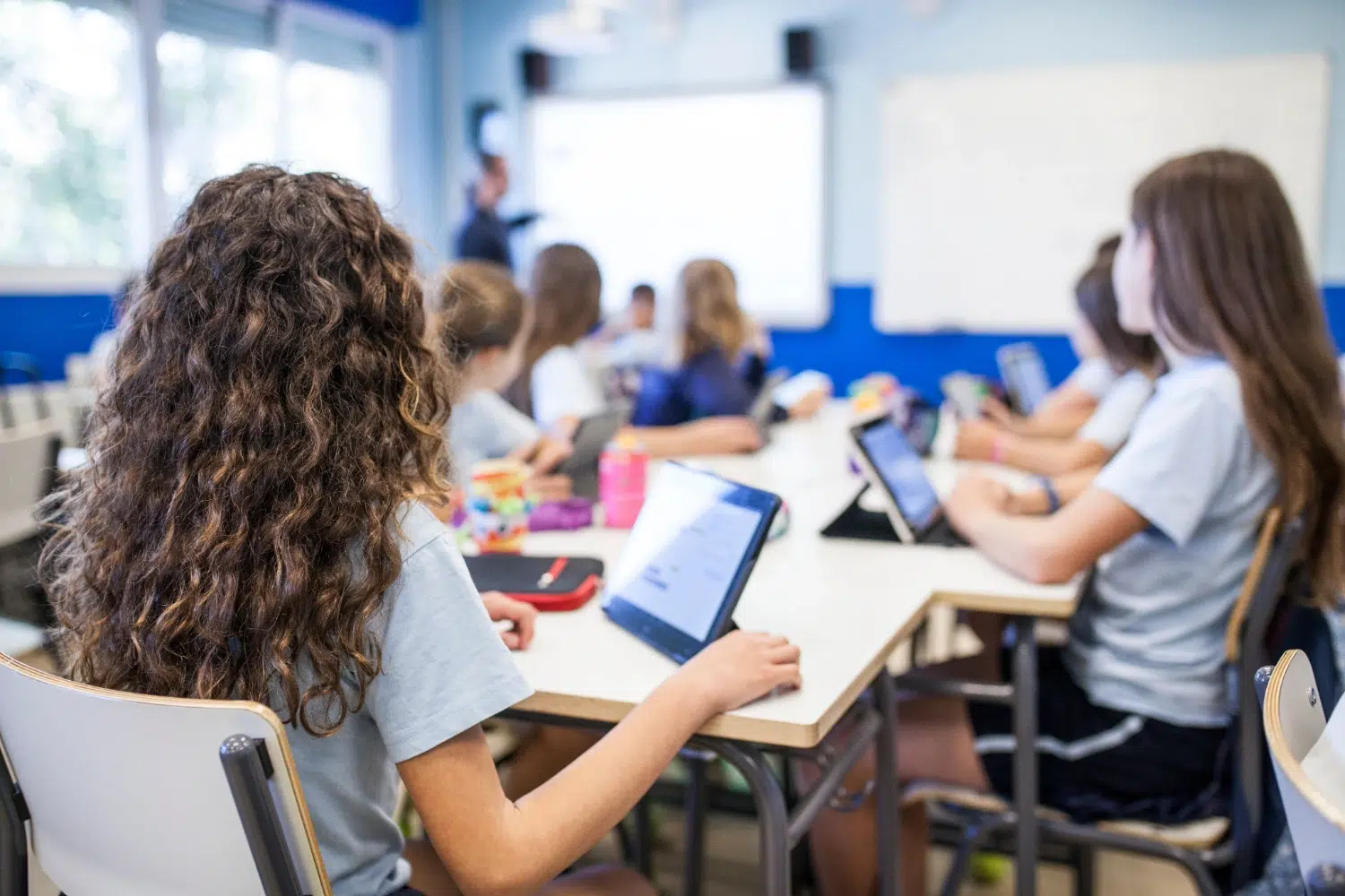 Students in a bright classroom sit at a long table using tablets while a teacher writes on a whiteboard at the front.