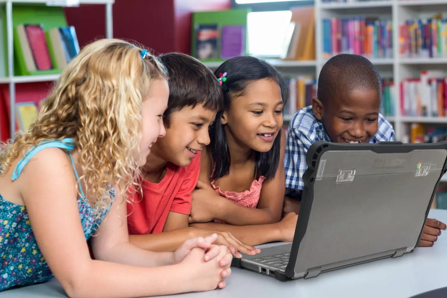 Four children of diverse backgrounds lean in to look at a rugged laptop on a classroom table, smiling as they explore together in a library.