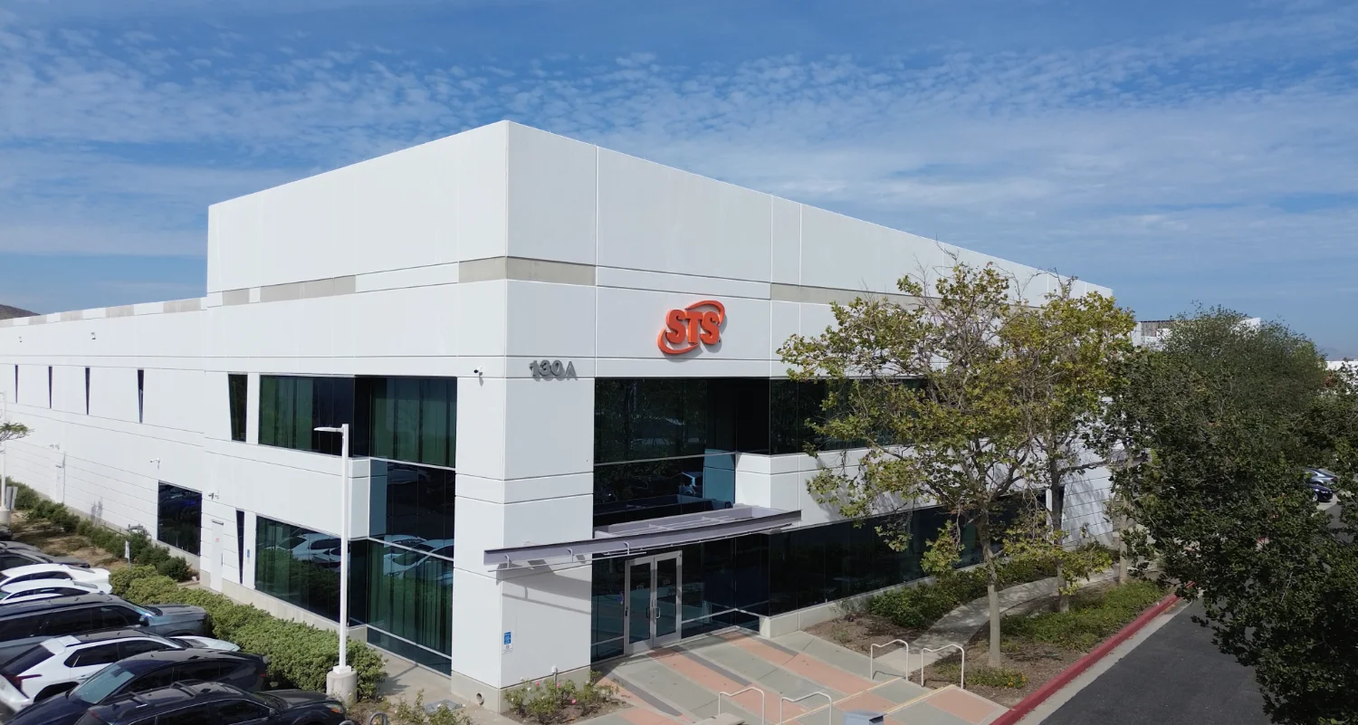 Modern white office building with STS logo on the façade, glass windows, and a row of parked cars nearby on a sunny day.