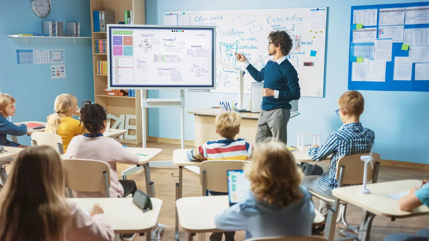 Teacher explains concepts at a whiteboard with a large digital display while students sit at desks in a bright classroom.