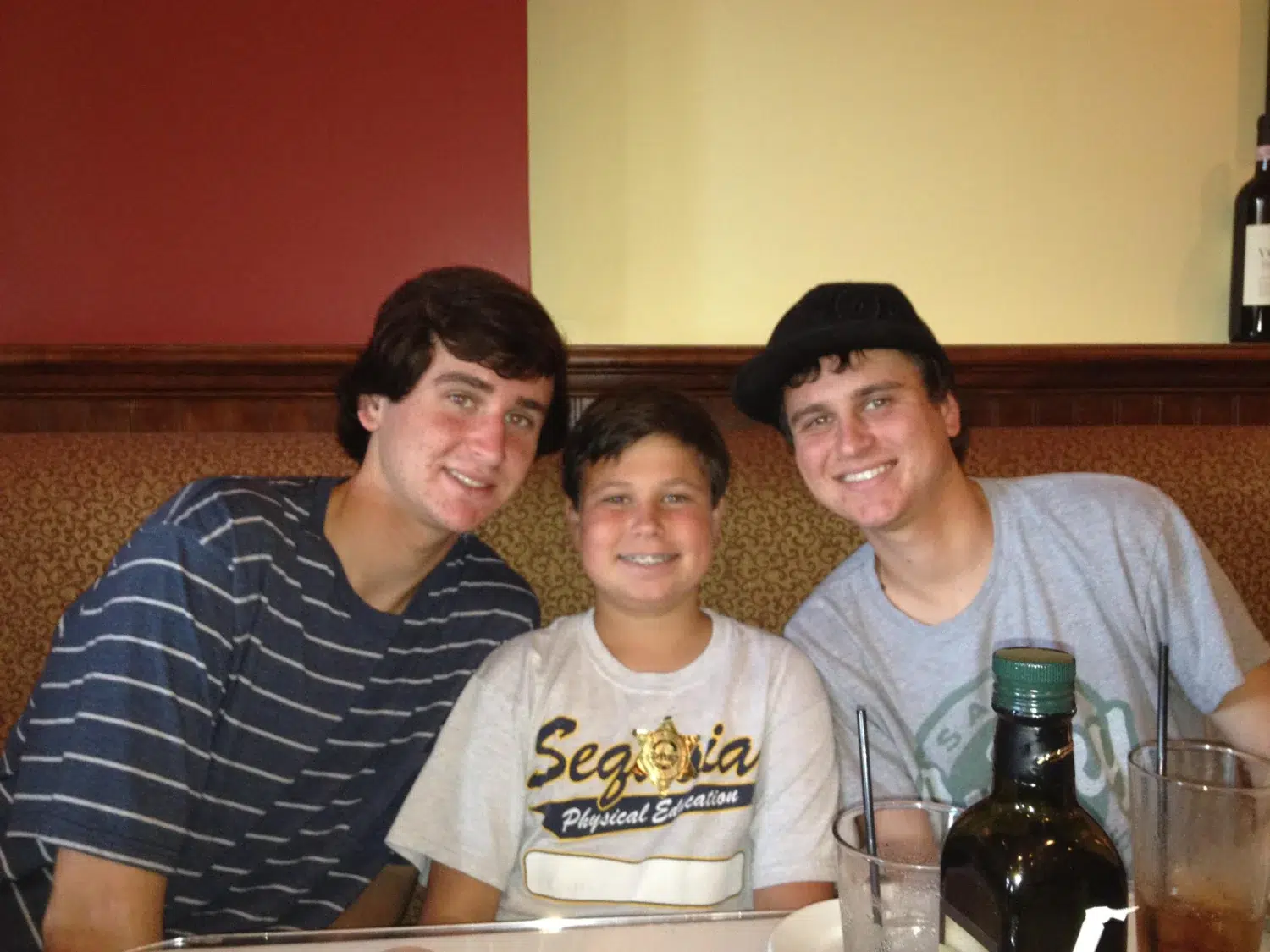 Three people sit close together in a restaurant booth and smile at the camera: left is a teen in a blue striped shirt, middle a younger boy in a light gray shirt, right a teen in a gray shirt wearing a black hat; drinks and a bottle are on the table behind them.
