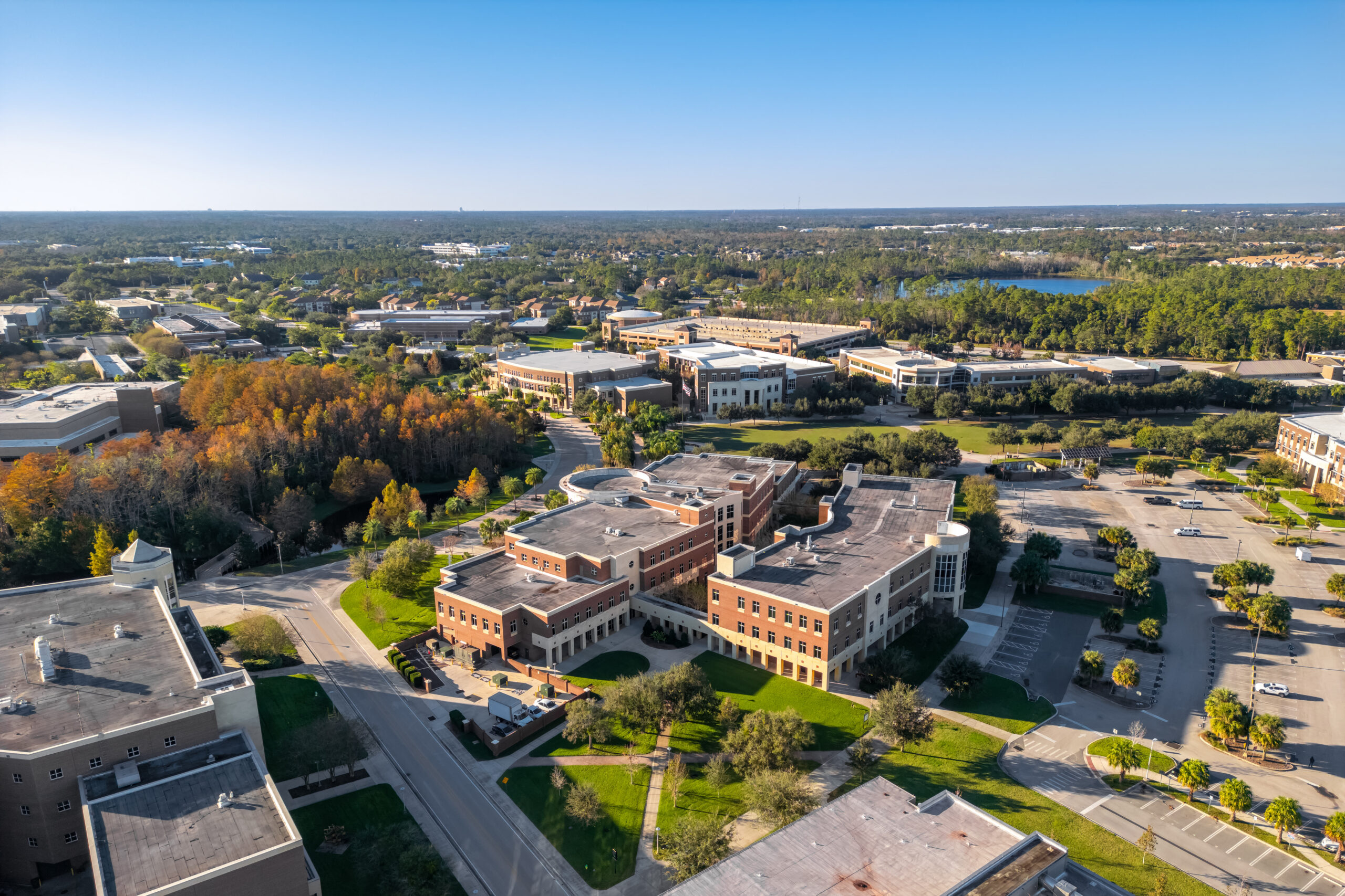 Aerial view of a university campus with brick academic buildings, parking lots, green lawns, and a lake visible in the distance on a sunny day.