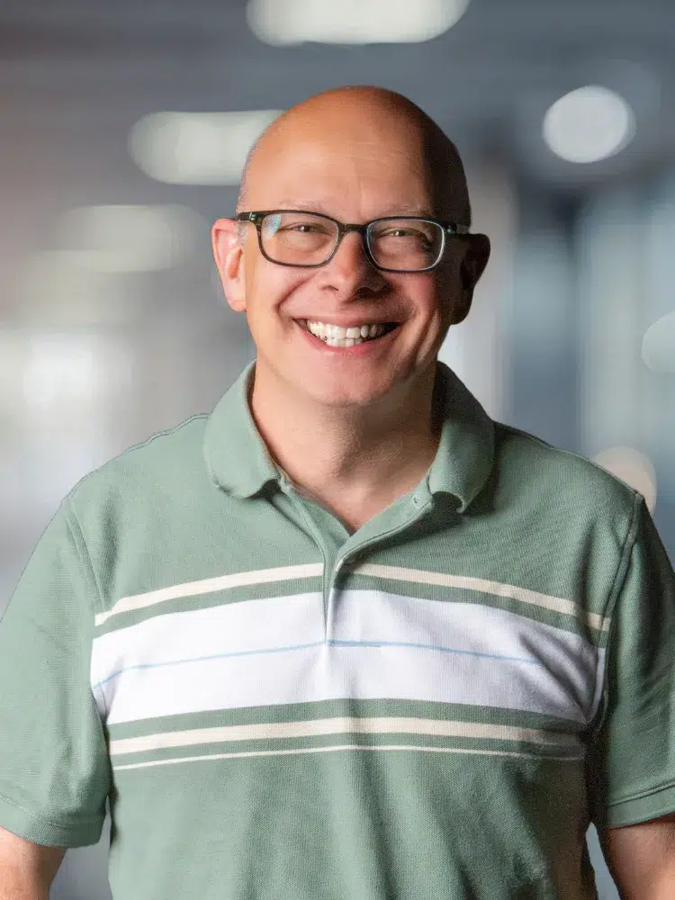Smiling man with glasses in a green striped polo shirt, office background.