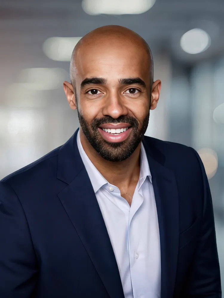 Professional headshot of a smiling man with a shaved head and beard, wearing a navy blazer and light shirt in a blurred office background.