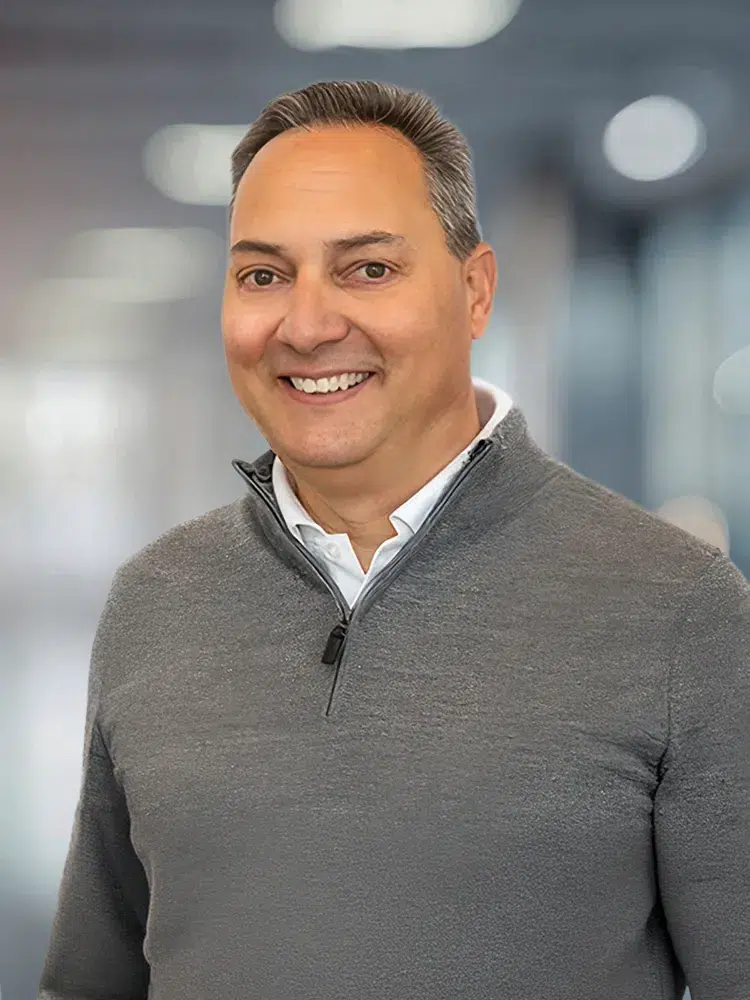 Smiling man in a gray quarter-zip sweater standing in a bright office setting.