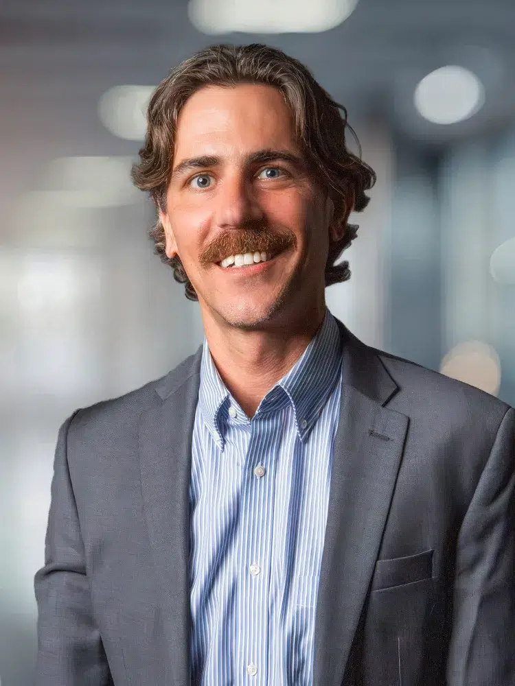 Professional man in a gray blazer and blue striped shirt, smiling in a modern office setting.