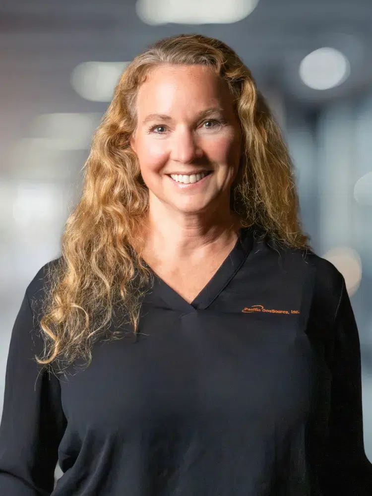 Professional woman with long curly blond hair smiling at the camera in a dark polo, office background blur behind her, logo on the shirt.