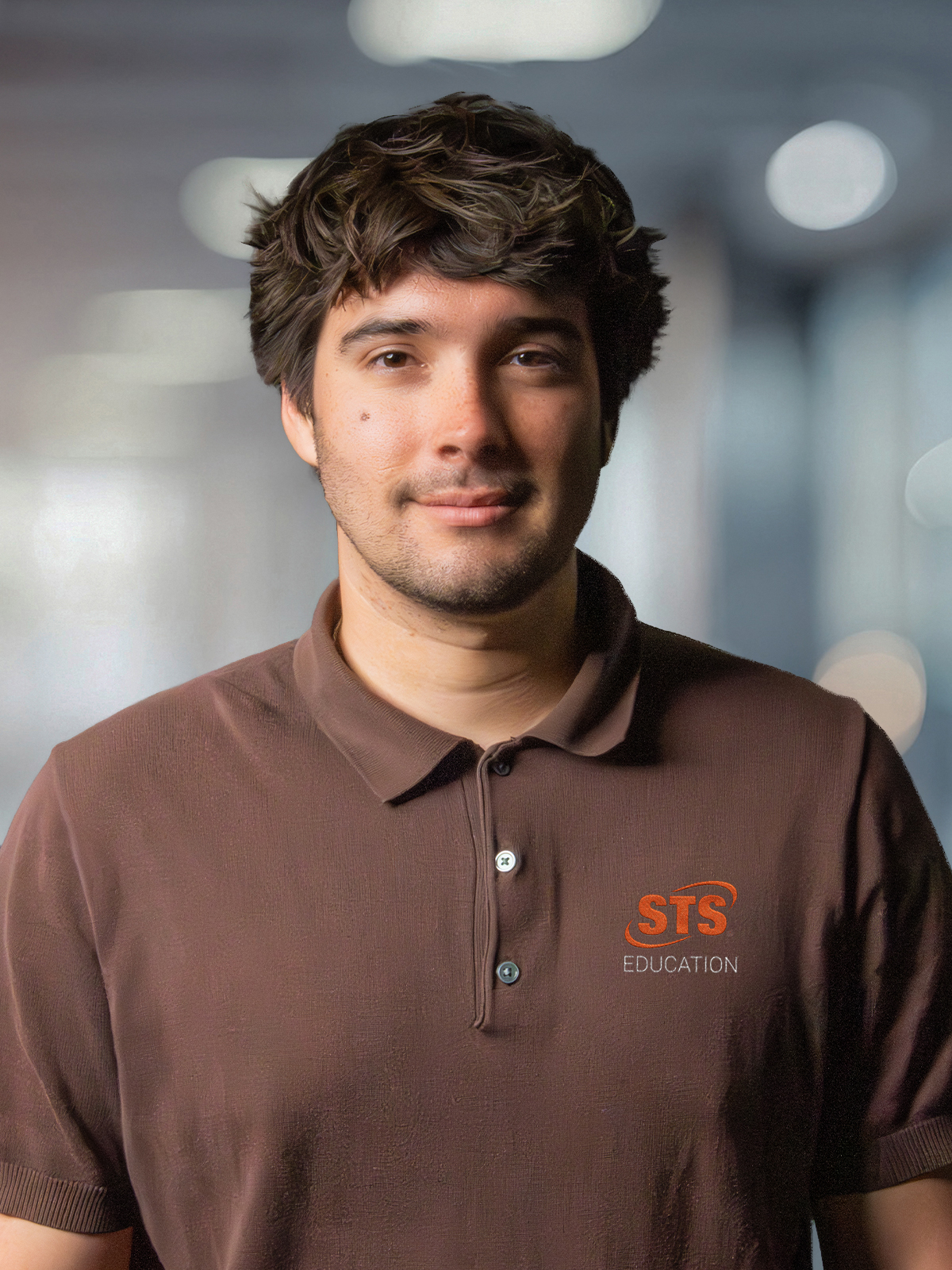 Portrait of a man with dark hair wearing a brown polo shirt featuring the sts education logo, against a blurred office background.