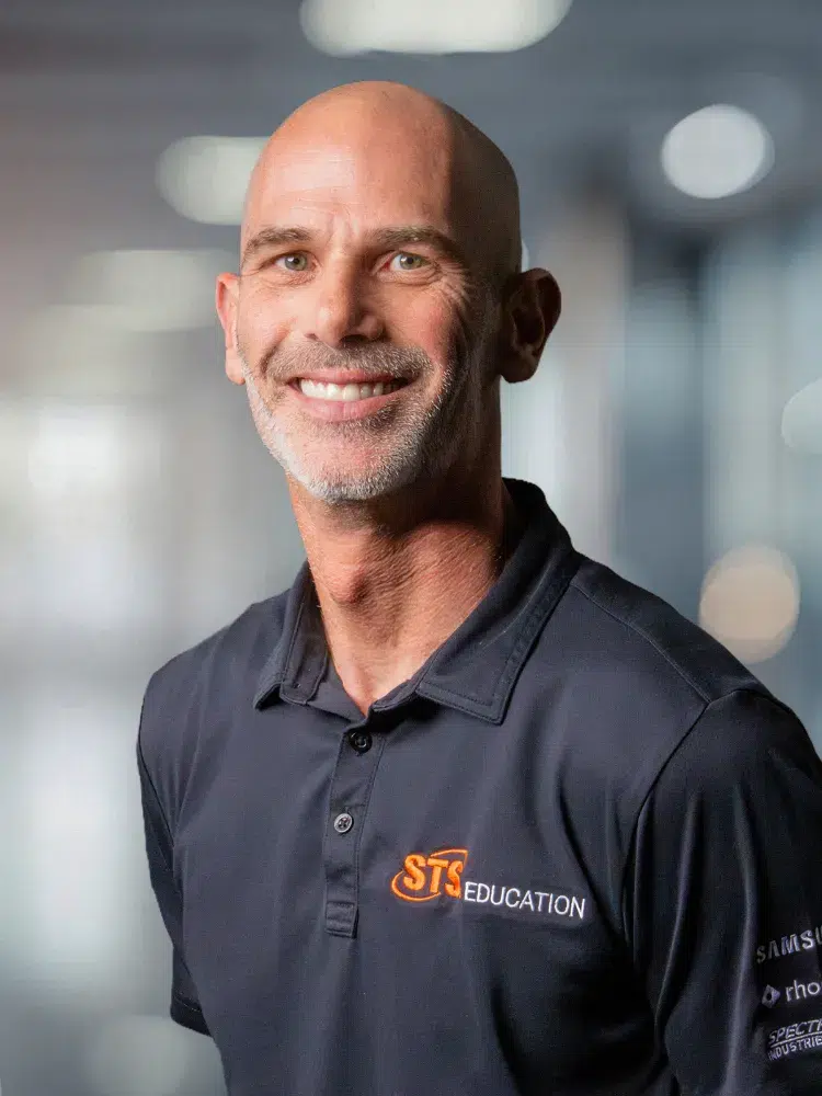 Smiling bald man with a gray beard wearing a dark polo shirt bearing the STS Education logo, in a blurred office background
