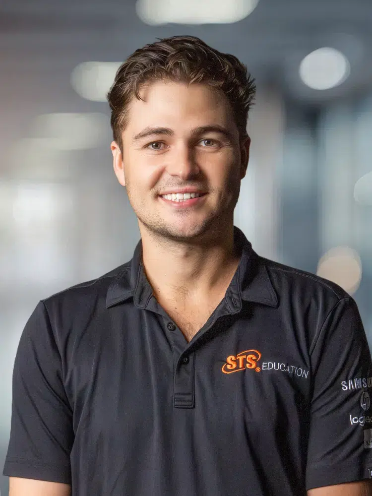 Smiling man in a black polo shirt with STS Education logo, standing in a softly blurred office hallway background.