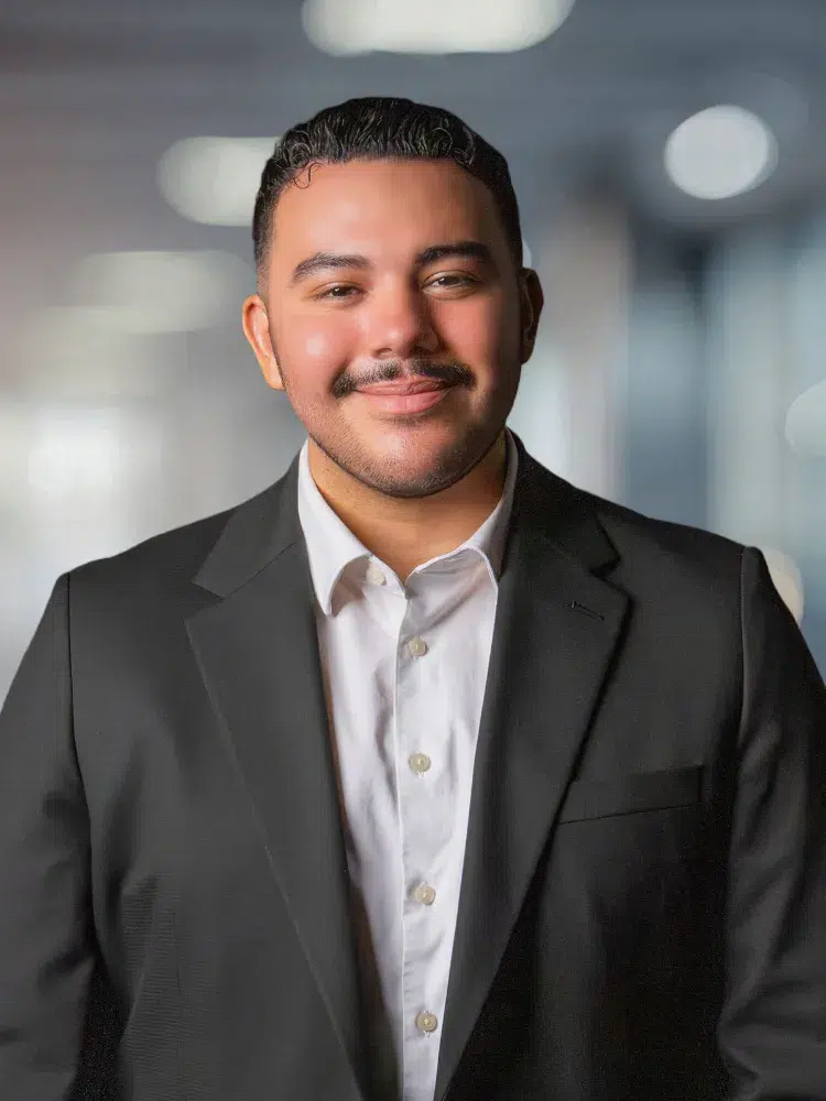 Portrait of a smiling man in a black suit and white shirt against a blurred office backdrop.