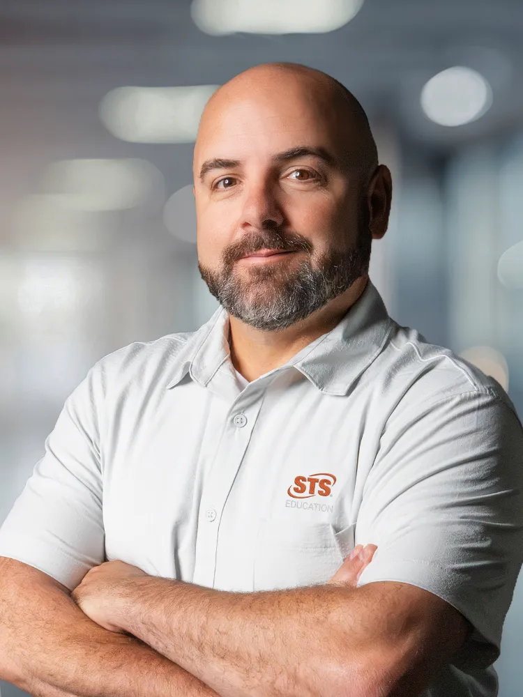 Portrait of a bald man with a beard, wearing a white polo shirt with the STS Education logo, arms crossed against a blurred office background.