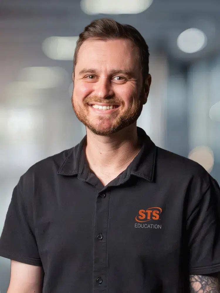 Smiling man in a black STS Education polo with a visible orange logo, standing in a bright office.