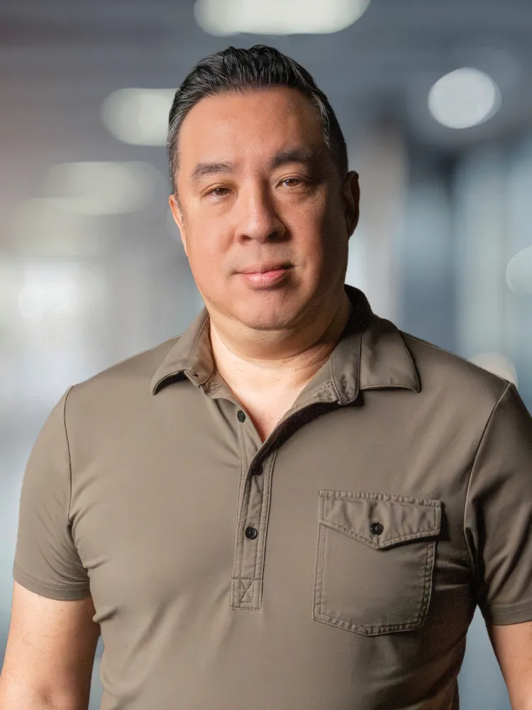 Portrait of a middle-aged man in a khaki polo shirt, standing in a softly blurred office background, looking at the camera.