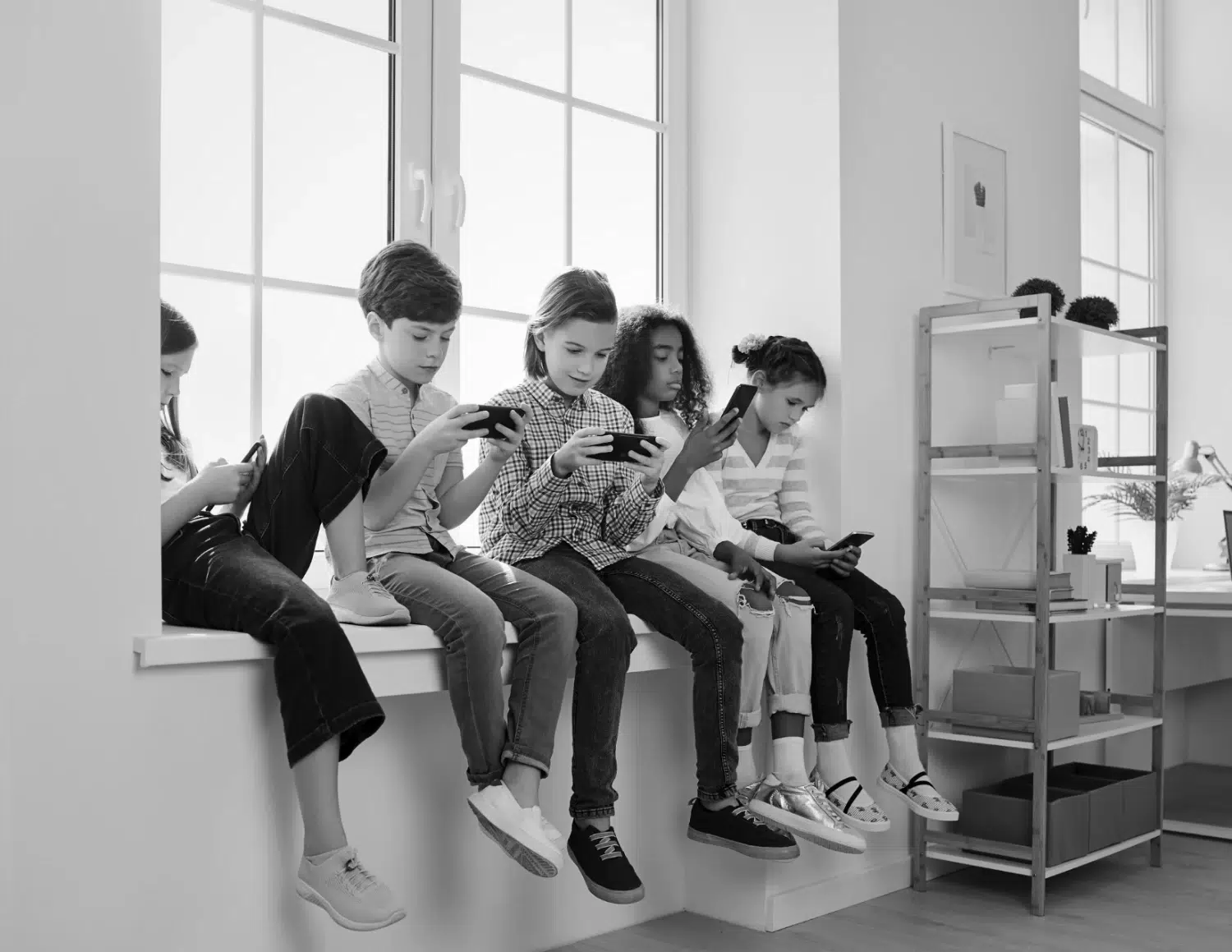 Five kids sit on a wide window ledge, each absorbed in handheld devices in a bright, modern room.