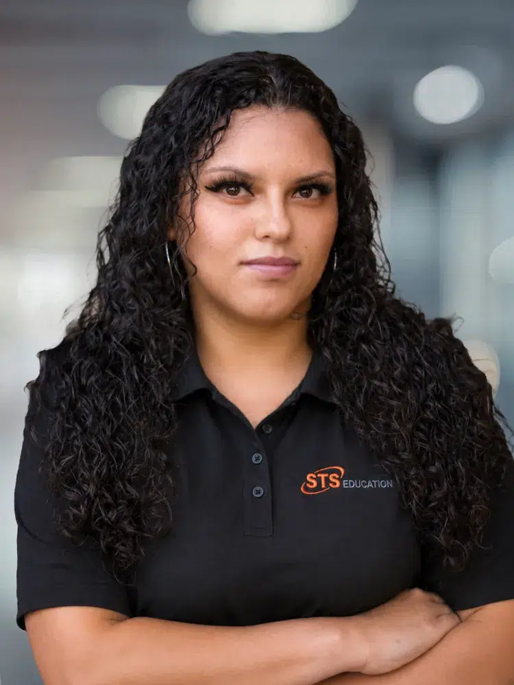 Portrait of a woman with long, curly black hair wearing a black polo featuring the STS Education logo, arms crossed, looking at the camera.