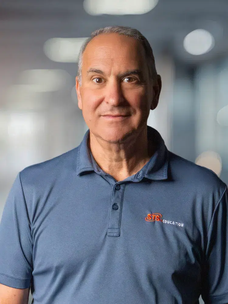 Portrait of a middle-aged man in a blue polo shirt with the orange 'STS Education' logo, standing in a bright office background.
