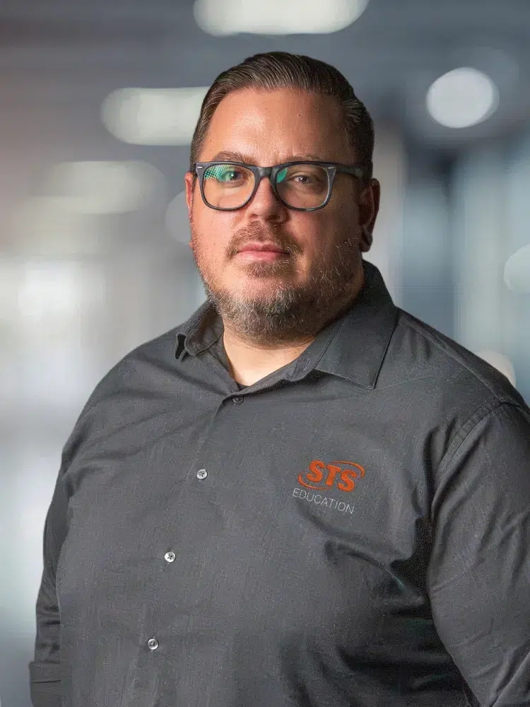 Portrait of a man with glasses wearing a gray STS Education shirt, in a blurred office background.