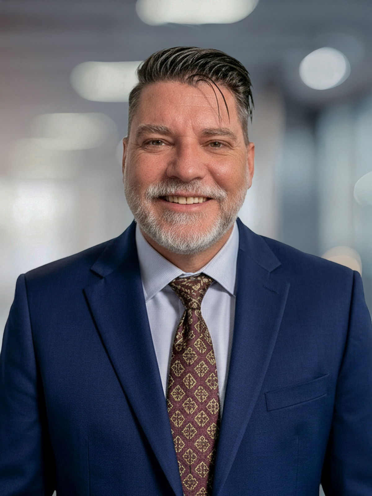 Professional head-and-shoulders portrait of a smiling man in a navy suit and patterned tie, office backdrop with soft bokeh lights.