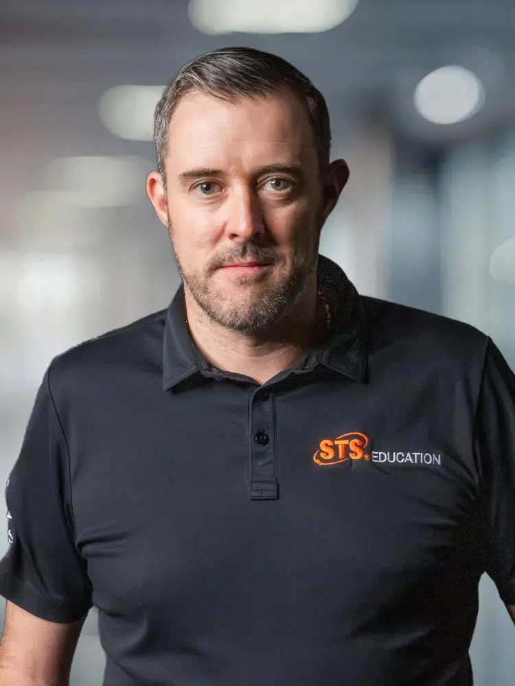 Portrait of a man in a black STS Education polo looking at the camera in a blurred office background.