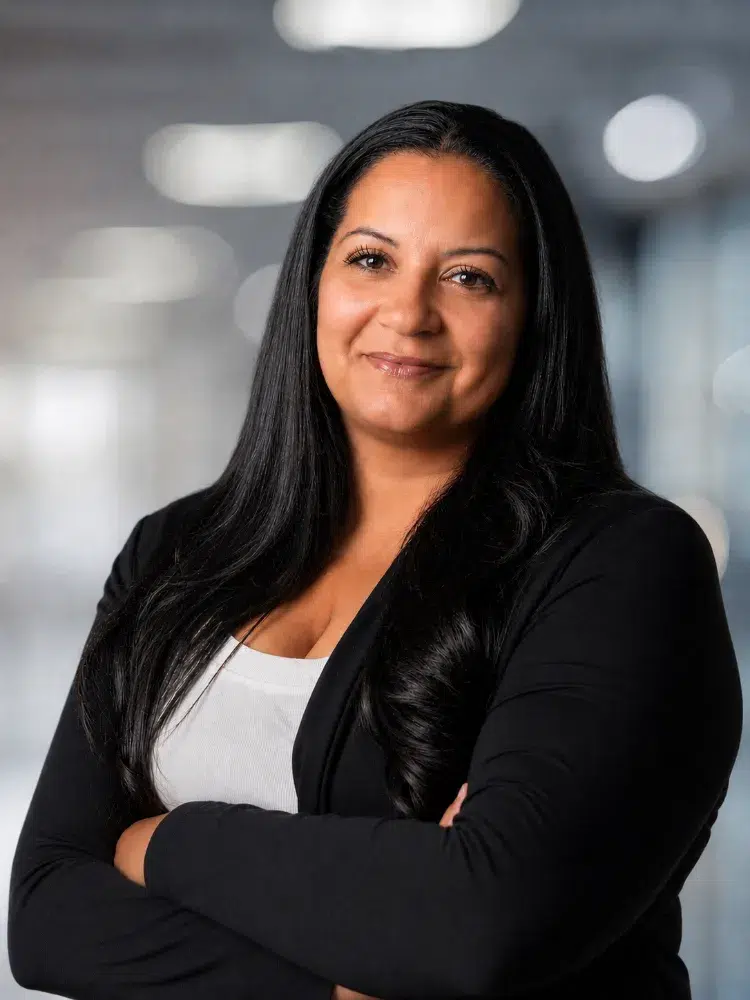 Portrait of a confident woman with long black hair, wearing a black blazer and white top, arms crossed in an office setting.