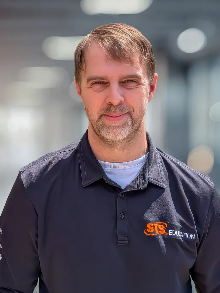 Man in a black STS Education polo shirt outdoors with a slight smile, facing the camera.