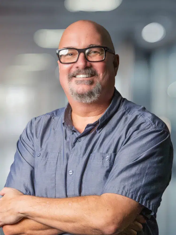 Smiling bald man with glasses and a gray goatee, wearing a blue button-down shirt with arms crossed in a blurred office setting.