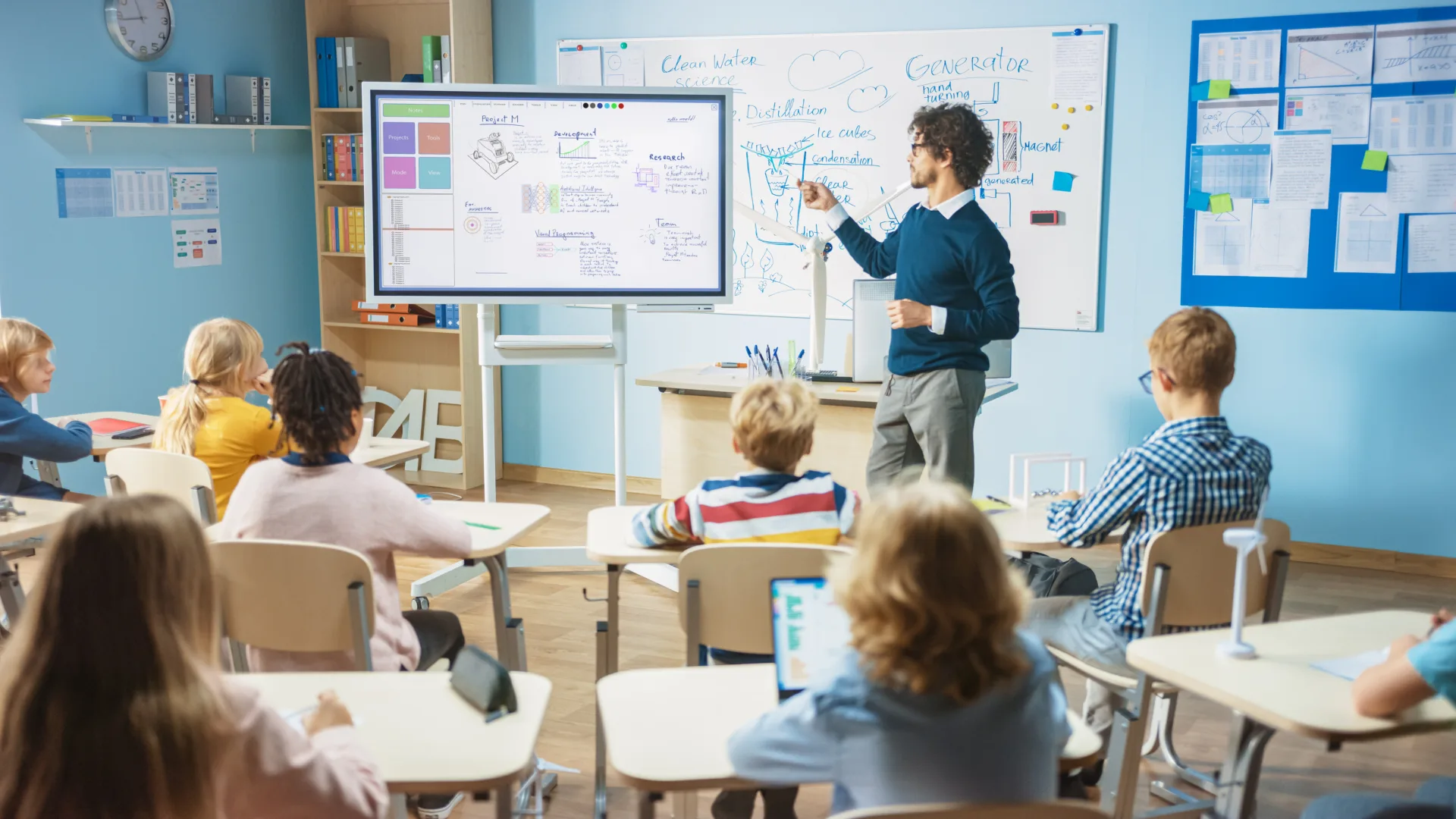 Teacher at whiteboard presenting a science lesson to students in a bright classroom with a large interactive screen nearby.