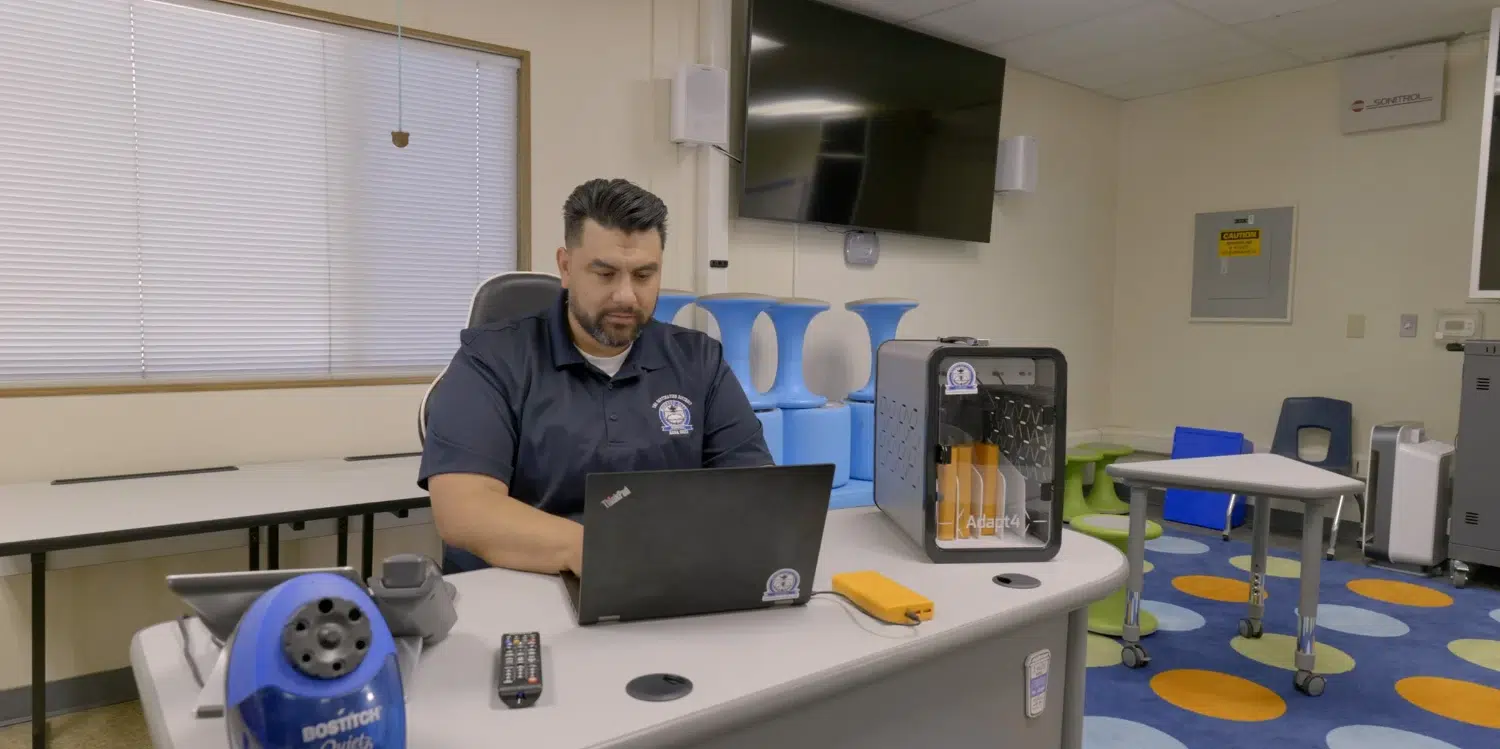 Man seated at a desk in a tech lab, using a laptop with a 3D printer beside him.