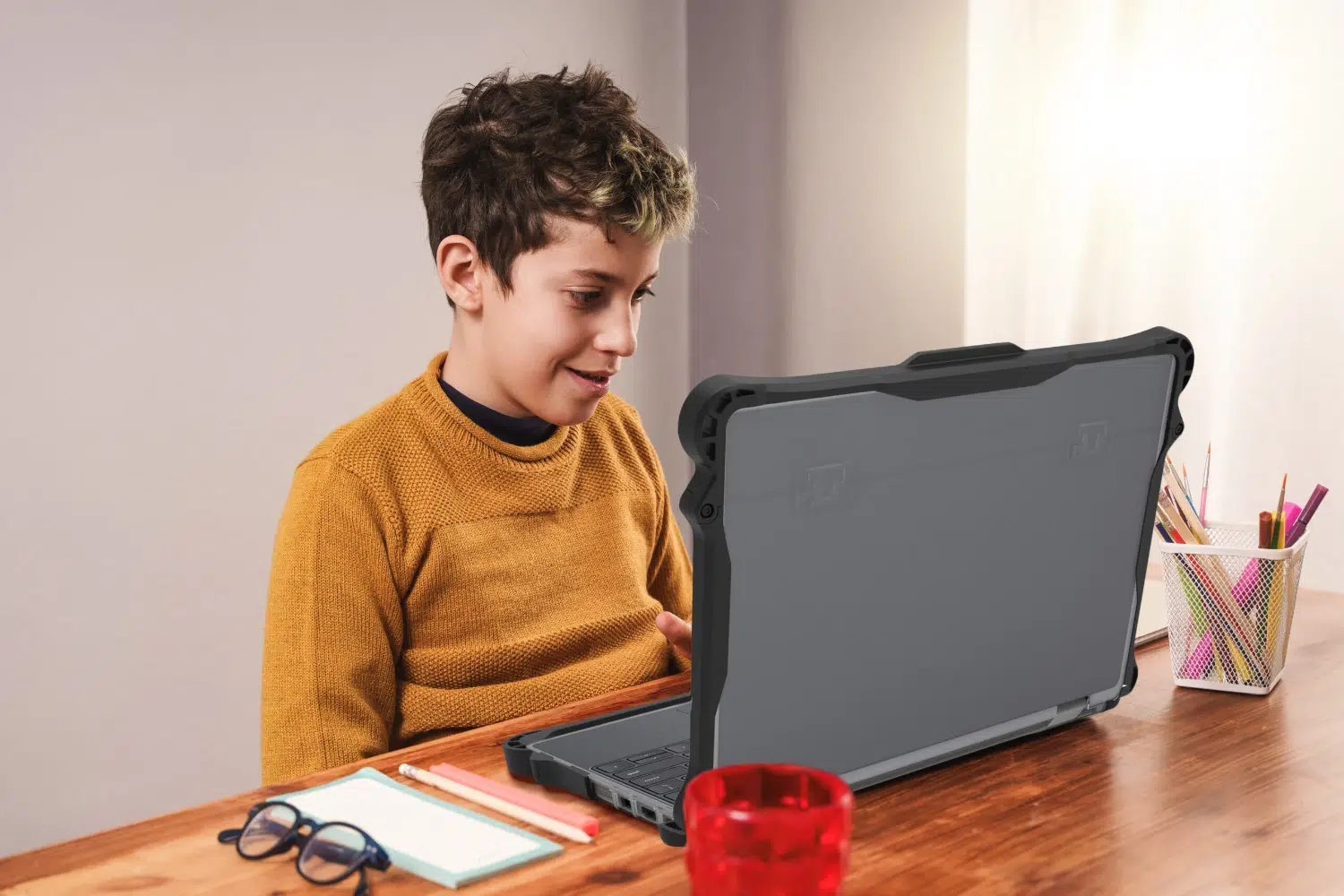 Young boy smiles while using a rugged laptop with a protective case on a wooden desk
