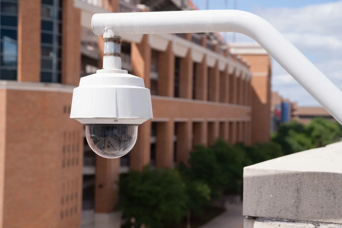 Outdoor security camera in a white dome mounted on a curved metal arm against a brick building backdrop on a campus roofline.
