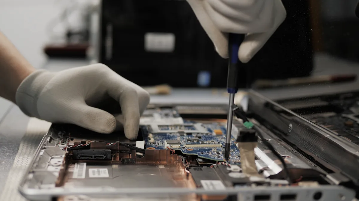 Technician in gloves repairing a laptop, using a screwdriver on the motherboard on a workbench.
