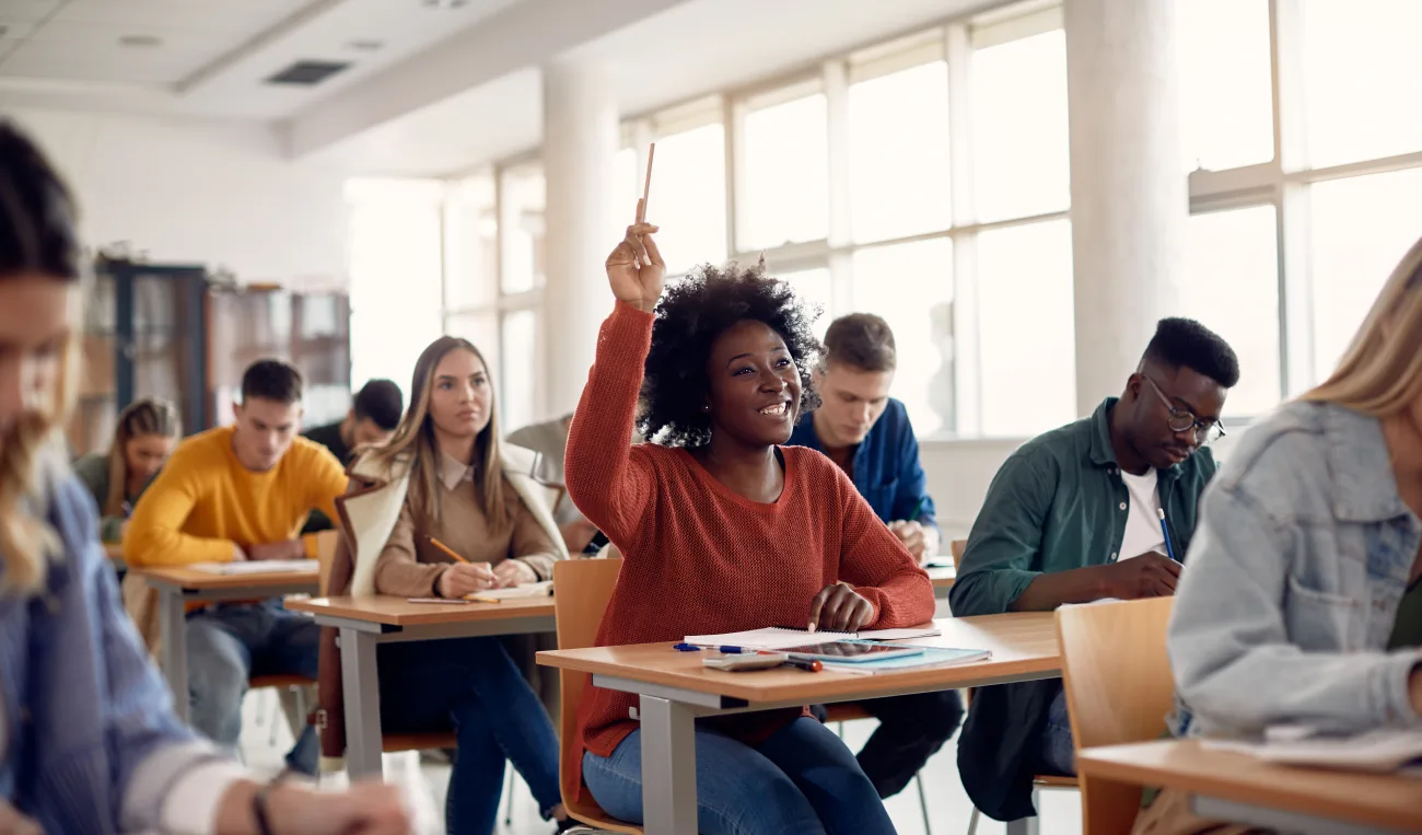 Black student in an orange sweater raises hand in a bright classroom while classmates study nearby.