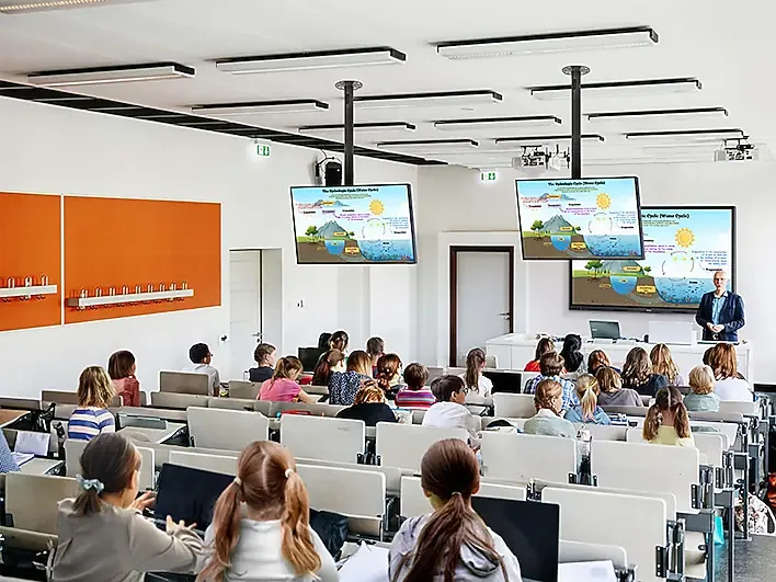 Lecture hall with students facing the front where three screens display slides as a presenter lectures from a podium on the right.