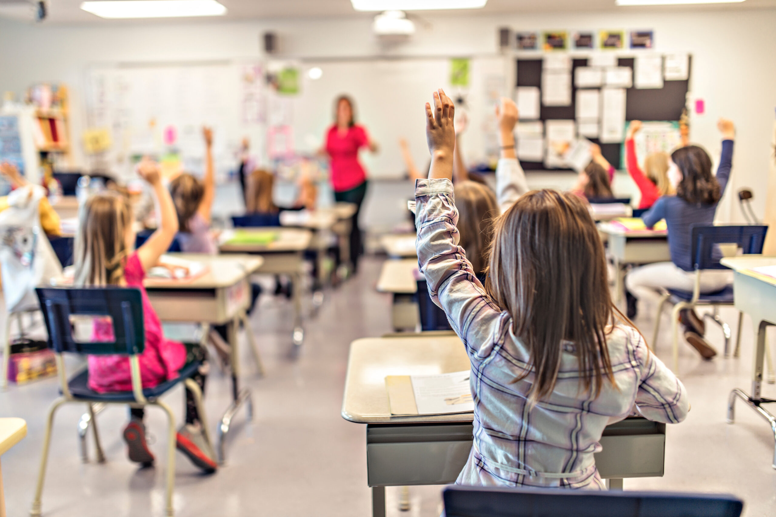 Students in a classroom raise hands as a teacher stands at the front near a whiteboard and projector.
