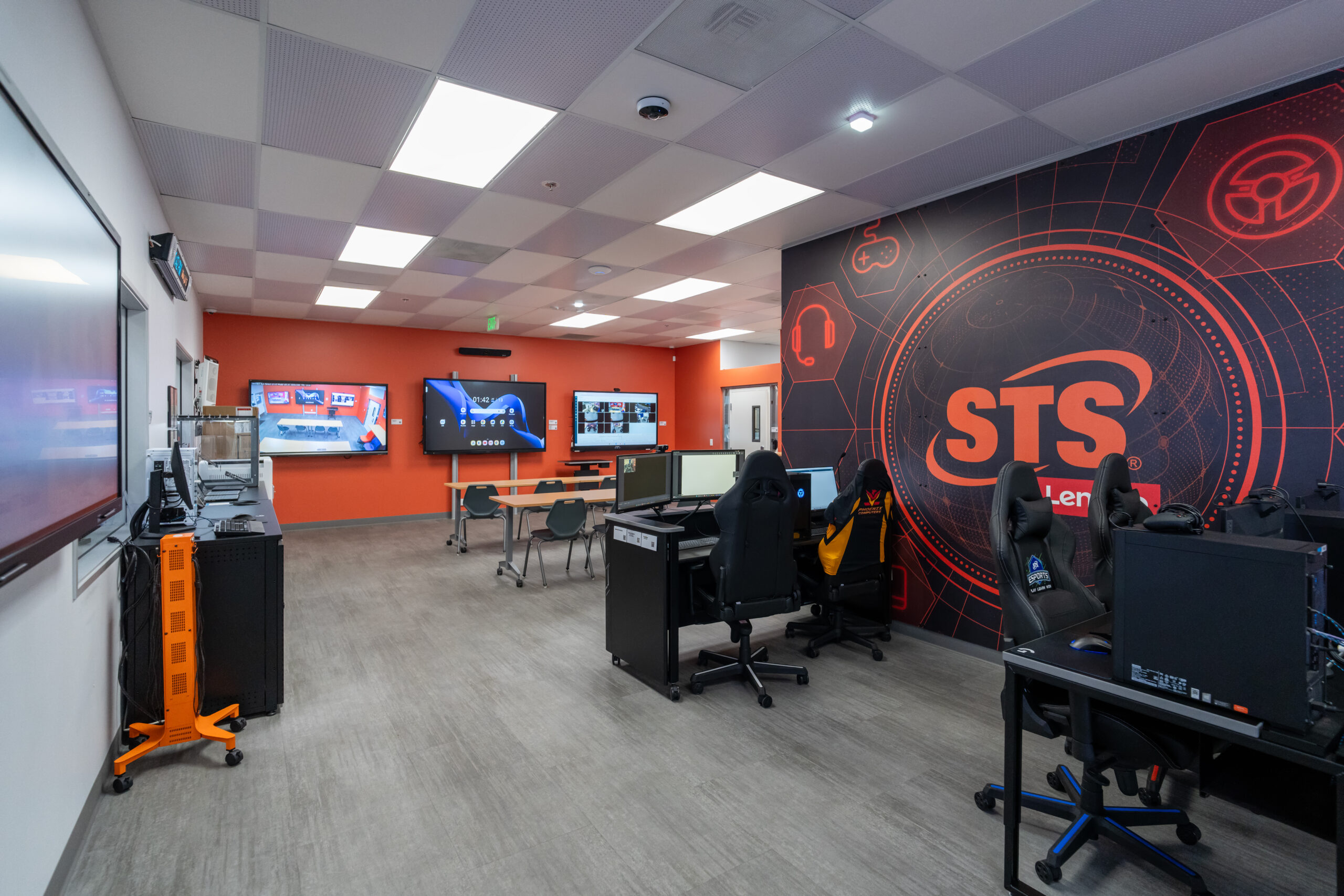 Esports training room with desks, multiple monitors, and black gaming chairs in front of a red wall mural with a large sts logo.