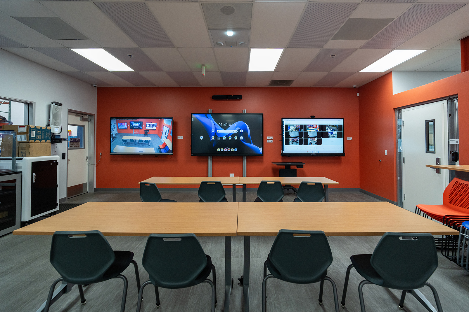 Classroom with an orange accent wall and three wall-mounted displays showing different content, plus wooden tables and teal chairs arranged for group work