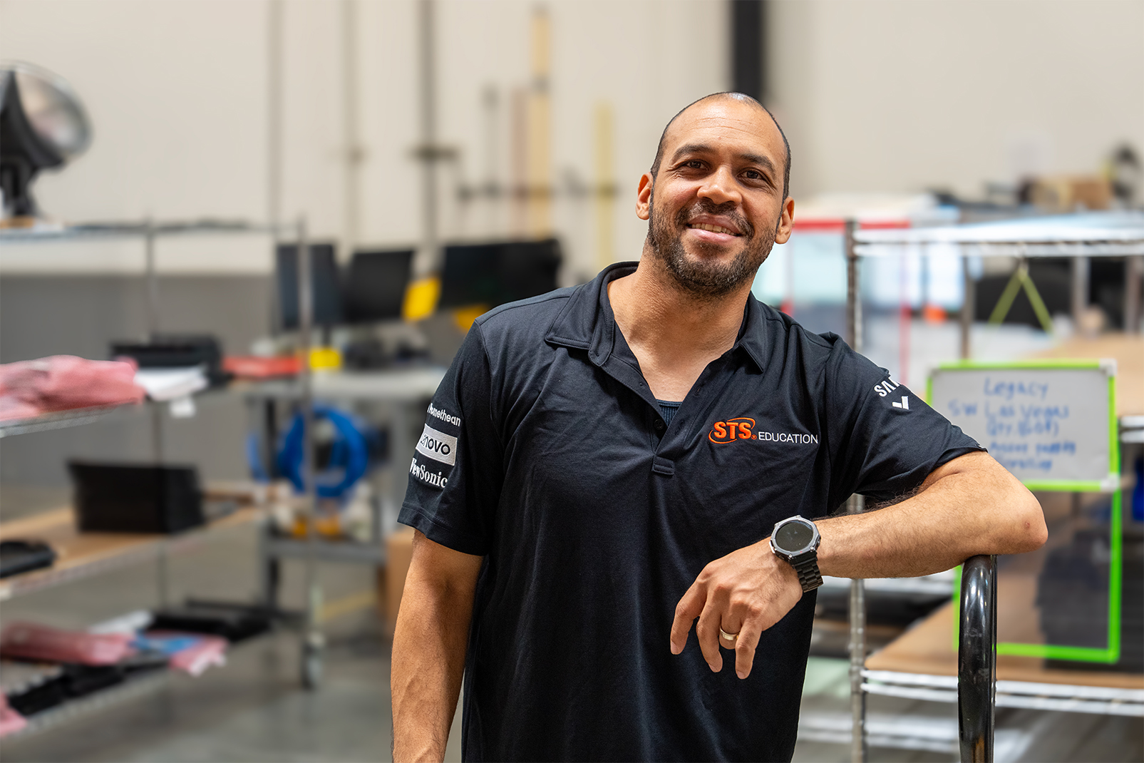 Smiling man in a black sts education polo stands in a workshop with shelves and equipment in the background.