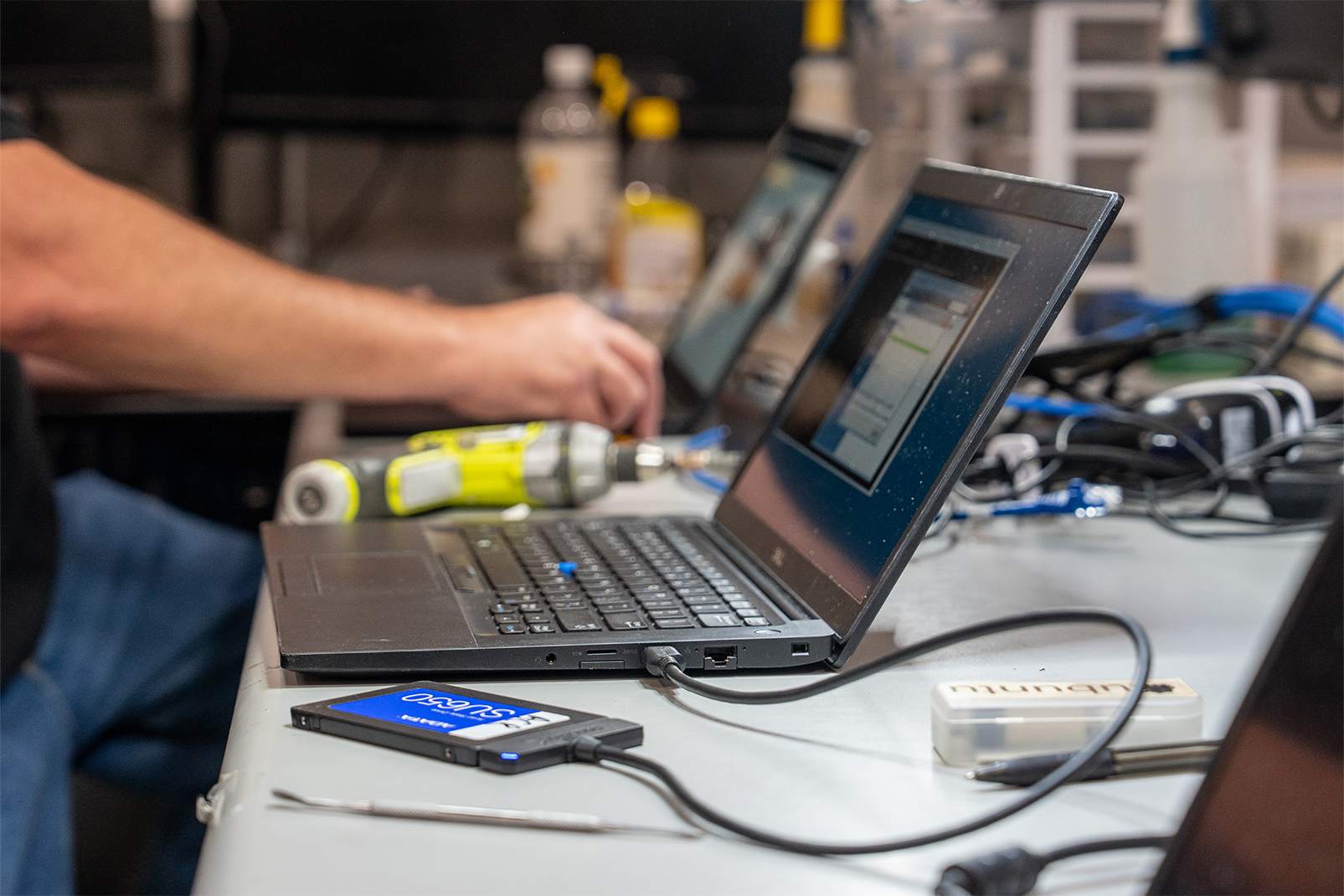 Person typing on a laptop at a cluttered lab bench with tools and cables nearby.