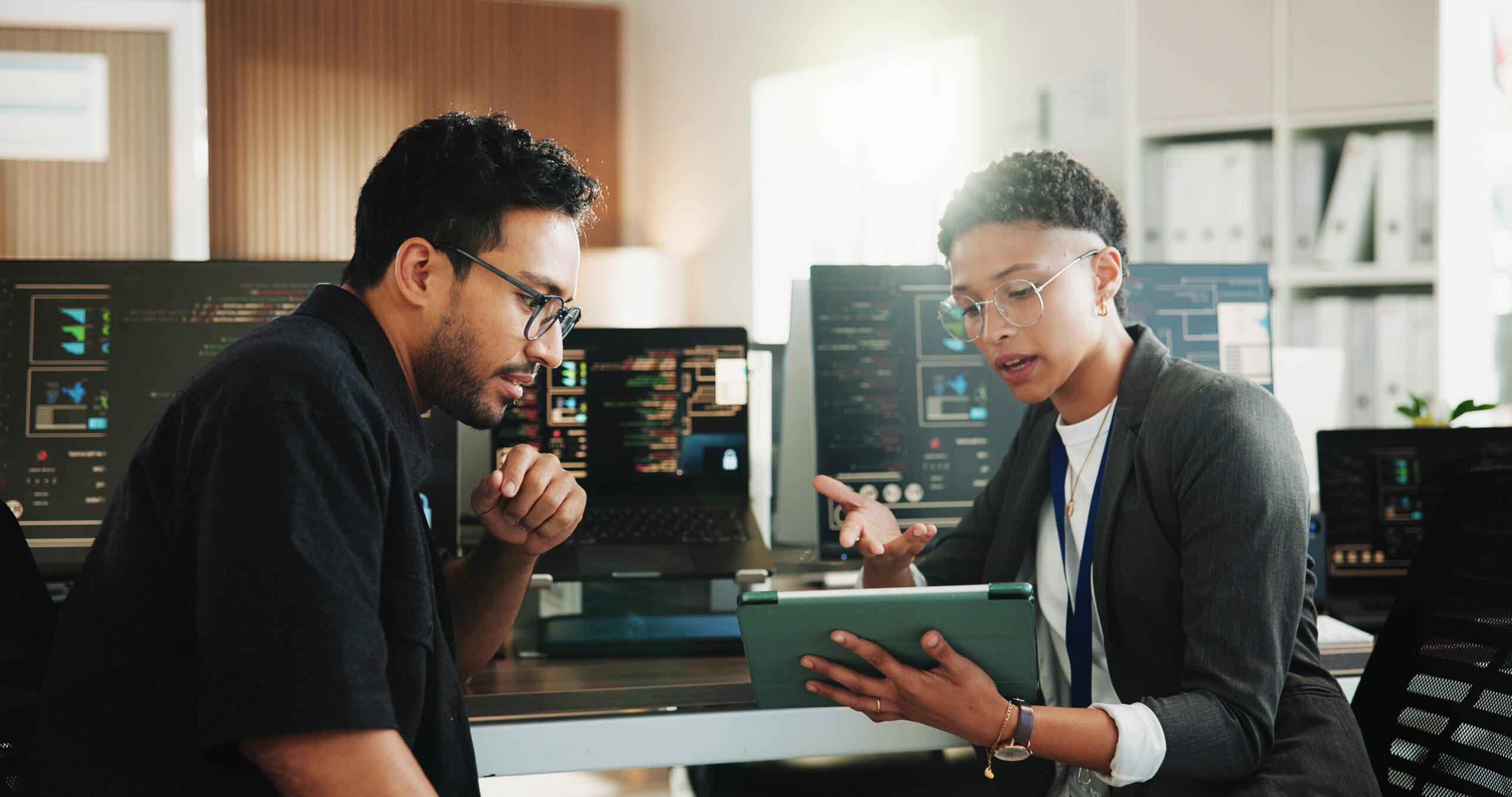 Two coworkers wearing glasses discuss data on a tablet in a modern office with multiple monitors behind them.
