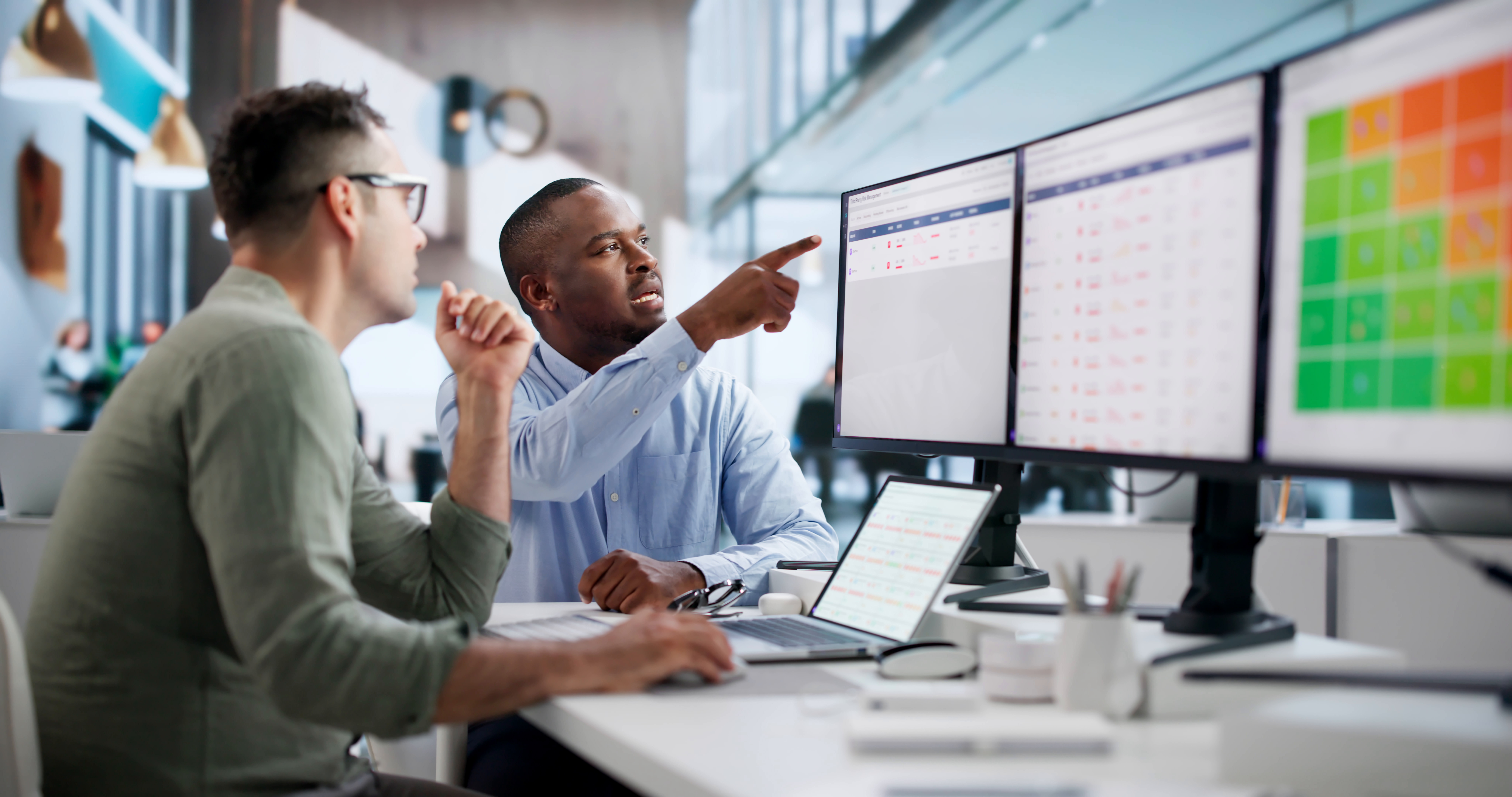 Two men in an office examine data on multiple monitors, one pointing at a dashboard while the other watches patiently.