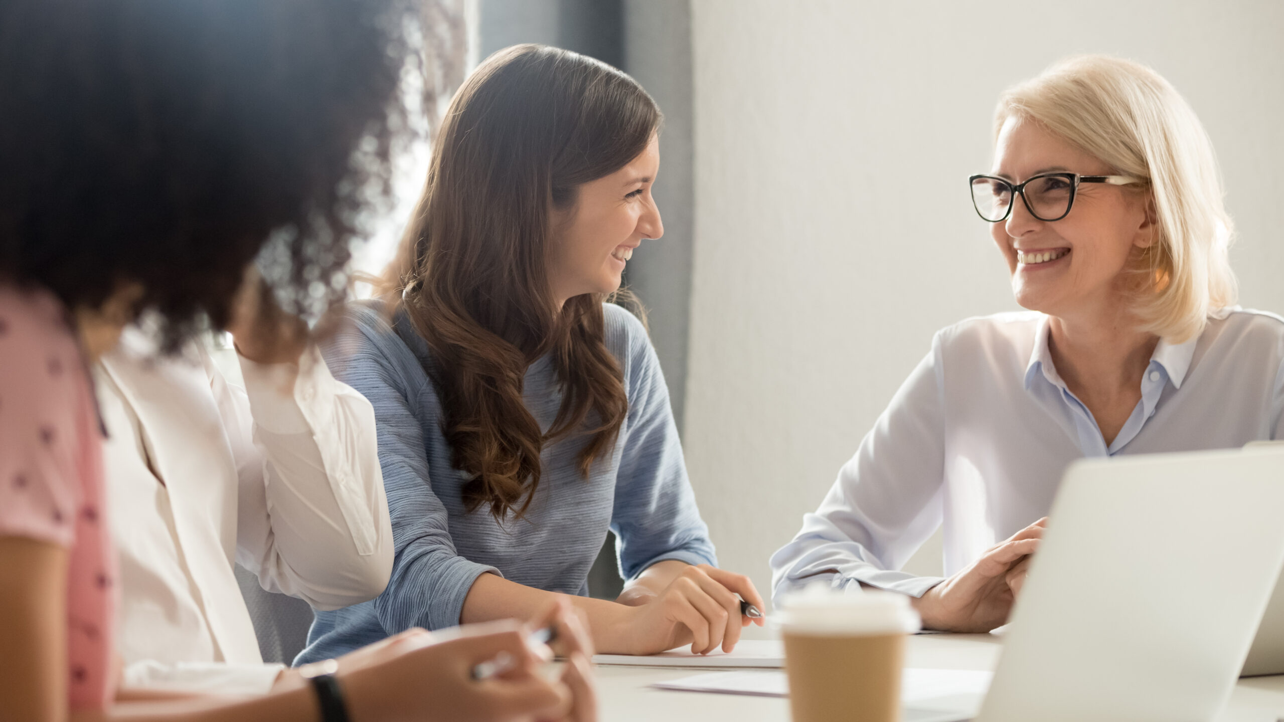 Team of coworkers at a table, smiling while discussing notes, with a laptop and a coffee cup visible.