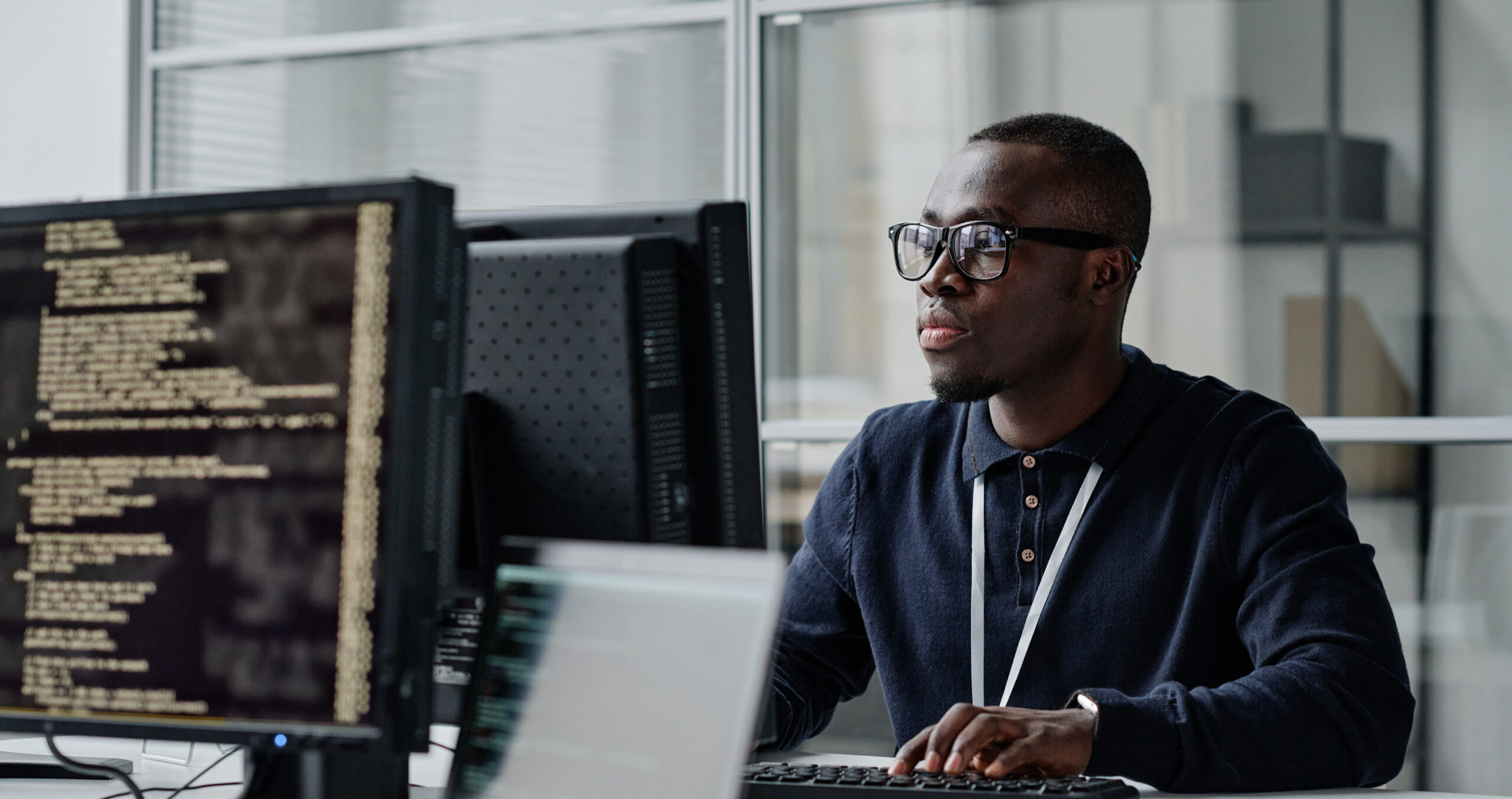 Man wearing glasses coding at a workstation with multiple monitors displaying code in an office setting.