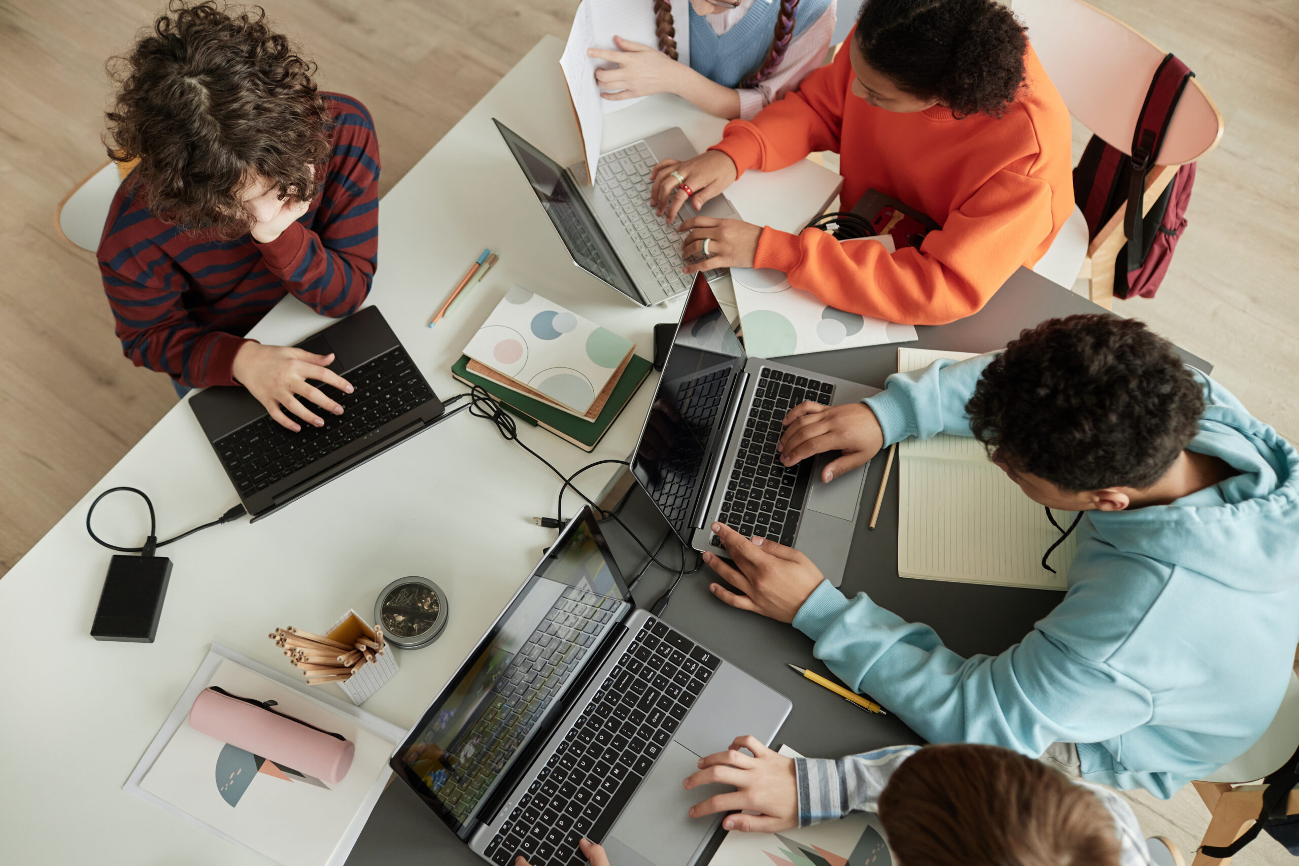 Overhead view of a group of students working on laptops around a table with notebooks, papers, and pencils for a collaborative study session.