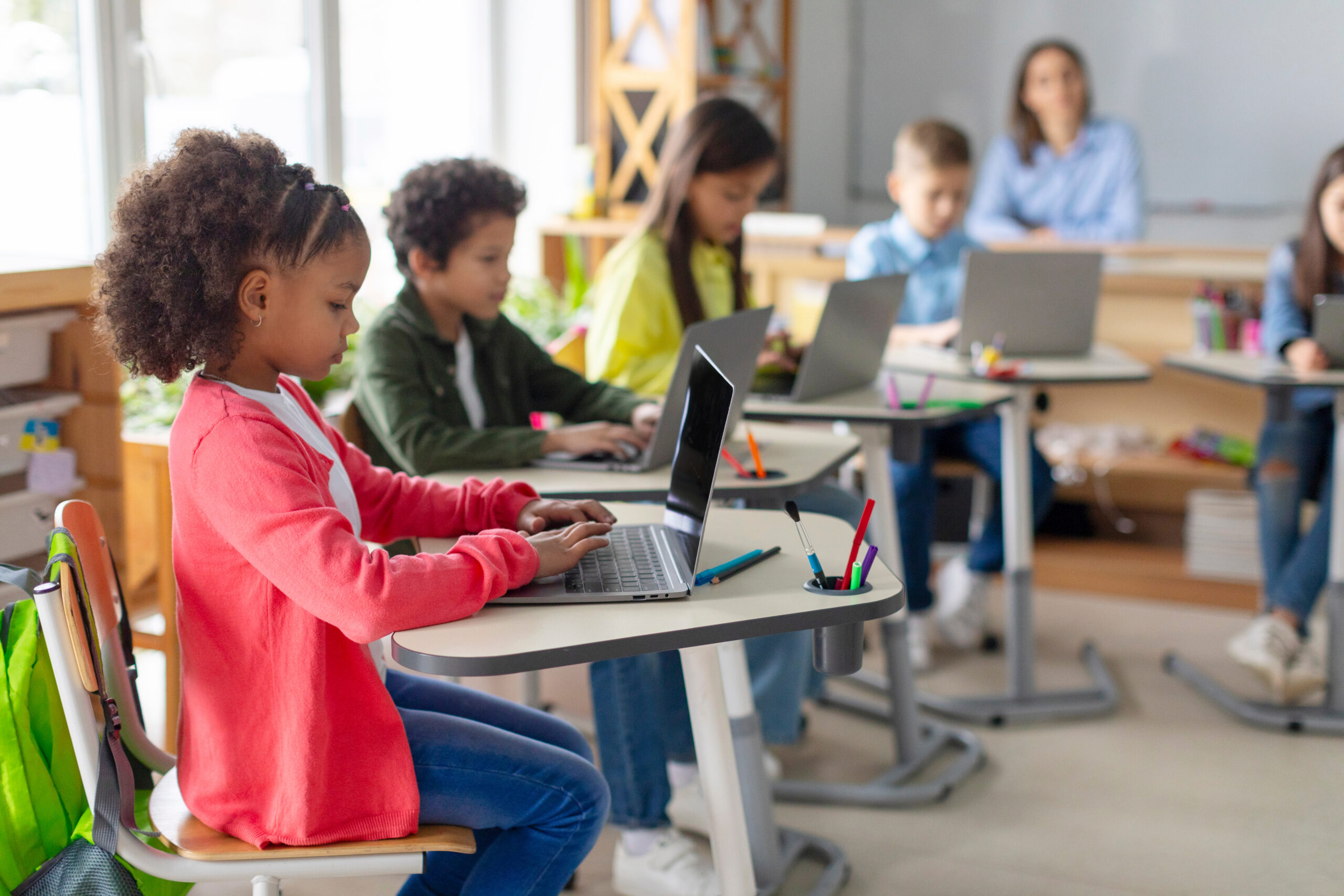 Young students sit at desks using laptops in a bright classroom, with a teacher in the background supervising back there.
