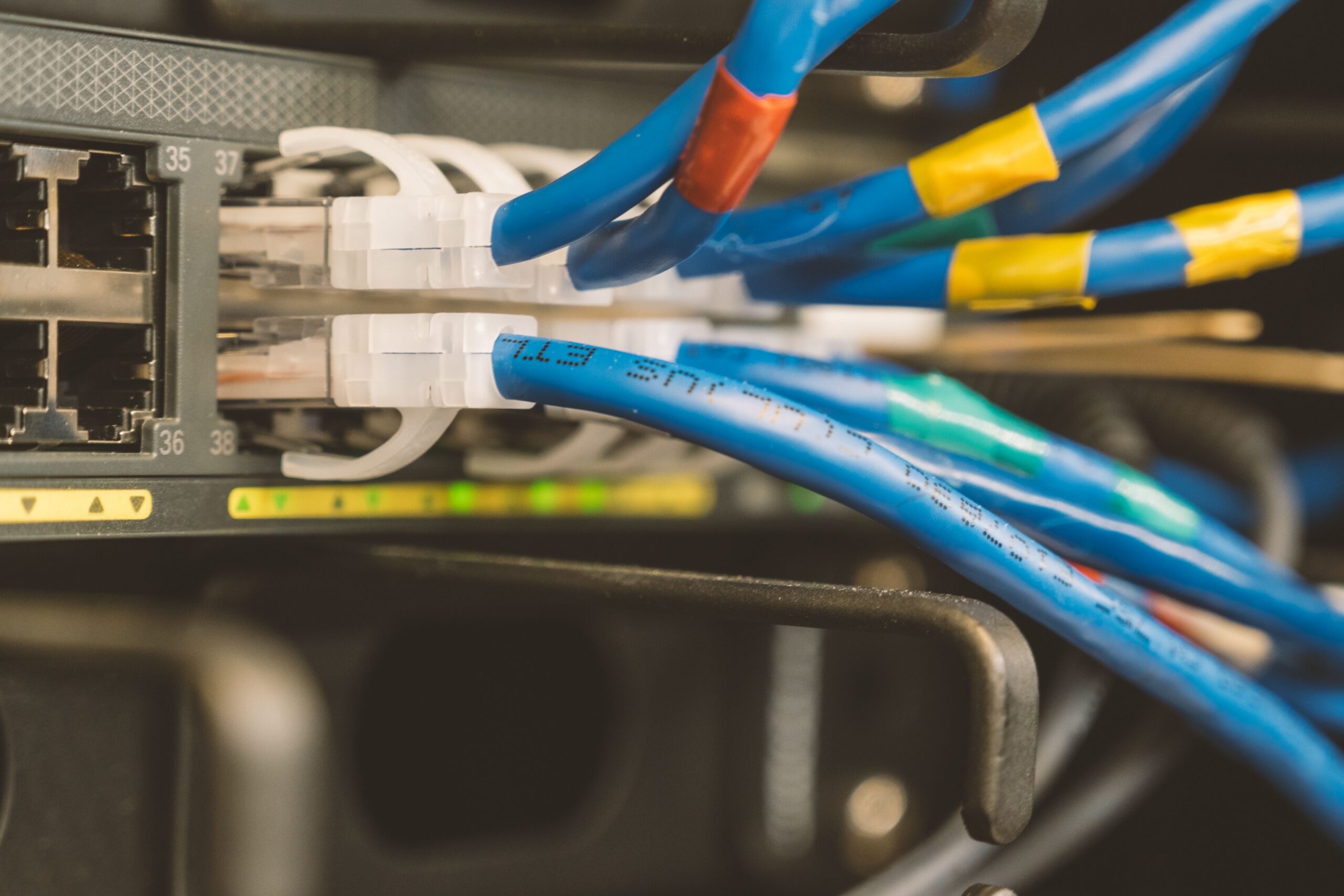 Close-up of blue ethernet cables with white connectors plugged into a network switch or patch panel in a server rack, with color-coded tape labels visible on the cables.